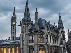 Gothic buildings with dominant towers and a clock tower, illuminated in the evening under a cloudy