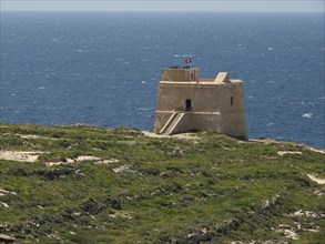 An old watchtower stands alone on a rocky coastline overlooking the vast sea, the island of Gozo