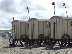 Changing cabins in the shape of carts stand on the beach, with red flags rising into the sky,