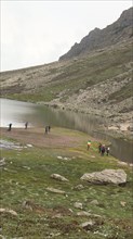 People near a small lake surrounded by rocky mountains, engaging in hiking on a cloudy day