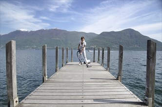 Happy and Elegant Woman Cleaning with a Vacuum Cleaner on a Pier over an Alpine Lake Maggiore with