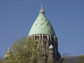 The upper part of a church tower with a greenish dome and a golden cross surrounded by tree tops,