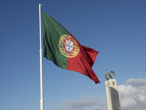 Portuguese flag flies high near a monument under a bright blue sky, Lisbon, Portugal, Europe