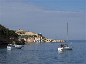 Sailing boats in front of a coastal landscape with cliffs and small houses under a partly cloudy