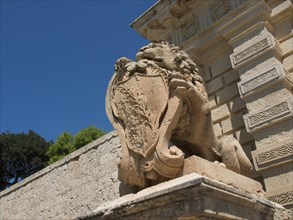 A stone lion sculpture holding a coat of arms, under a clear sky, the town of mdina on the island