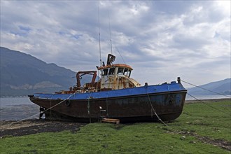 A rusty boat lies on the shore of a calm sea under a cloudy sky, surrounded by a green landscape,