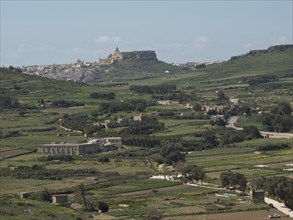 Wide landscape with fields and hills, in the background a settlement and a fort, the island of Gozo