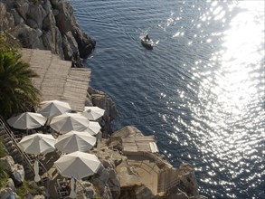View of the sea from the rock, with white parasols and a boat on the water, reflecting the sun's