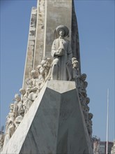 A stone monument with detailed sculptures of historical figures under a clear blue sky, Lisbon,