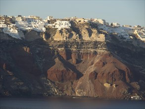White buildings characterise the top of the steep cliffs towering over the calm sea, brown volcanic