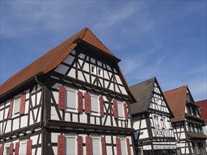 Three half-timbered houses with red shutters and tiled roofs, under a blue sky in the sun, historic