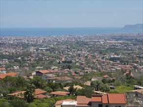 A wide panorama over a city with a multitude of houses, hills and the sea in the background,
