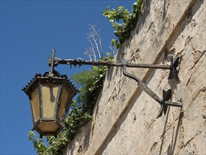 An old metal lantern on a stone wall, overgrown with plants under a blue sky, the town of mdina on
