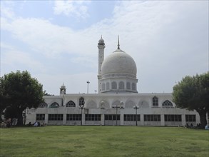 A white mosque with a large dome and minaret, surrounded by trees and grass under a cloudy sky