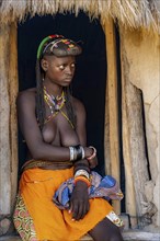 Hakaona woman with traditional kapapo hairstyle, sitting in the entrance of her mud hut, in the