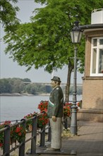 Statue of a man in uniform next to a walkway on the riverbank with lamps and blooming flowers,