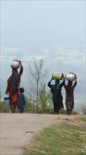 Group of people walking along a path carrying water containers in a serene rural area, wearing