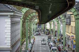 A suspension railway runs above a busy city street with cars and buildings in Wuppertal Vohwinkel