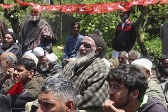 Group of people seated outdoors under flags, attentively listening, with trees in the background,