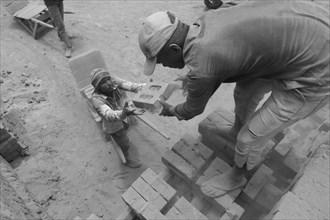 Child handing a brick to a man in a dusty construction site in black and white, Jammu and Kashmir,