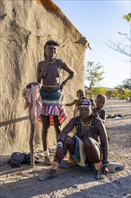 Two Hakaona woman with traditional kapapo hairstyle, with children, in front of their mud hut, in