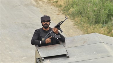 An armed soldier holding a rifle, emerging from a vehicle hatch on a road during the day,