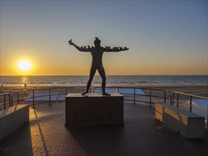 A statue in front of a sunset on the beach, surrounded by a railing, sunset in many colours on the