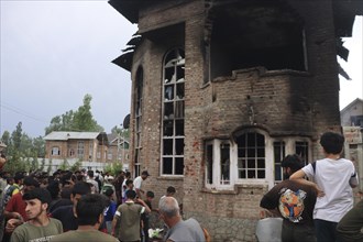 A crowd of people stands around a damaged and burnt multi-story brick building, observing the