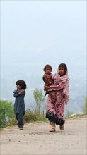 Children walking along a rural road, with one child carrying another, wearing traditional clothing,