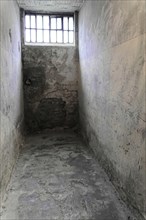 Cramped cell with weathered walls and a small barred window. Deserted and cold surroundings,