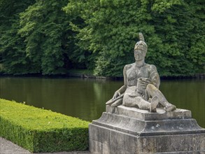 Stone sculpture of a seated man next to a pond, surrounded by lush greenery and trees, old red
