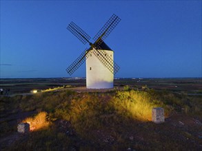 An illuminated windmill on a field hill at dusk under a clear blue sky, aerial view, Alcazar de San