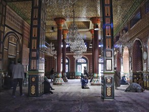 Ornate and colorful mosque interior with chandeliers, intricate designs on pillars, and people