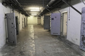 Long prison corridor with open metal doors and grey walls. Gloomy lighting, cold atmosphere,