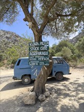 Tourist humorous funny sign on tree behind old Volkswagen VW Bus Bulli in front of entrance of