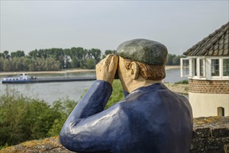 A statue of a sailor with binoculars looking at a river and a boat, next to a building, rhine