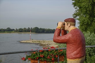 Man statue with binoculars on the riverside, flowers in the foreground and trees in the background,