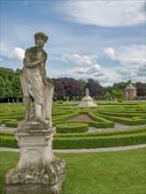 Stone sculpture of a woman in a well-kept garden with decorative hedges and surrounded by trees,