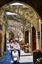 Man riding a scooter through an old, sunny street with stone arches and historic buildings, Old