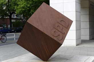 Red-brown cube with SPD inscription in front of a modern building on a street with a bicycle in the