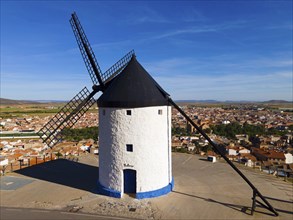 A traditional windmill with white and blue colours in front of a village and a wide sky, aerial
