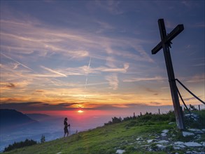 Young slim woman with hiking poles next to a summit cross at sunset, cirrus clouds in the sky,