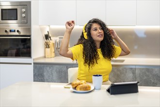 Latin young woman enjoying music while having breakfast sitting on the kitchen at home
