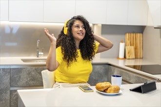 Happy latin young woman listening music with headphones sitting in the kitchen eating breakfast at