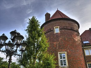 Historic brick tower with windows, flanked by a tree and antique lanterns under a partly cloudy
