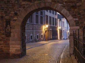 A stone archway offers a view of an illuminated cobblestone street with historic buildings at