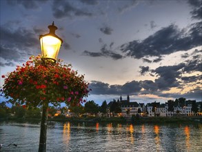 Illuminated street lamp with flowers at dusk on the river bank, reflected light in the water,