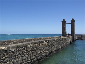 A stone footbridge leads to the sea with a Tor tor in the foreground and blue water all around,