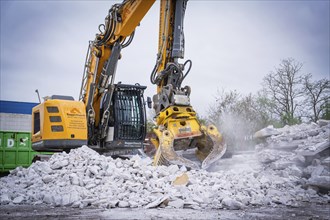 Large yellow excavator with metal grab shredding rubble on a construction site, trees in the