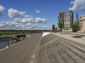 Riverbank with modern buildings in the background and a promenade, under a sky with small clouds,
